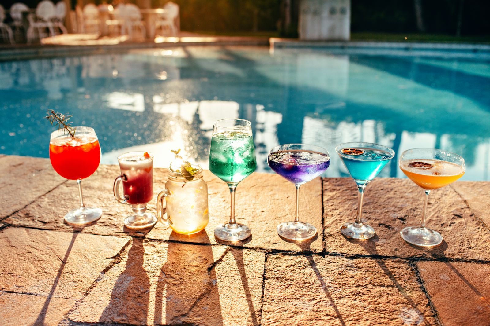 A row of colorful cocktails in different glasses on the edge of a pool in warm sunlight, the drinks behind every cruise drink package question