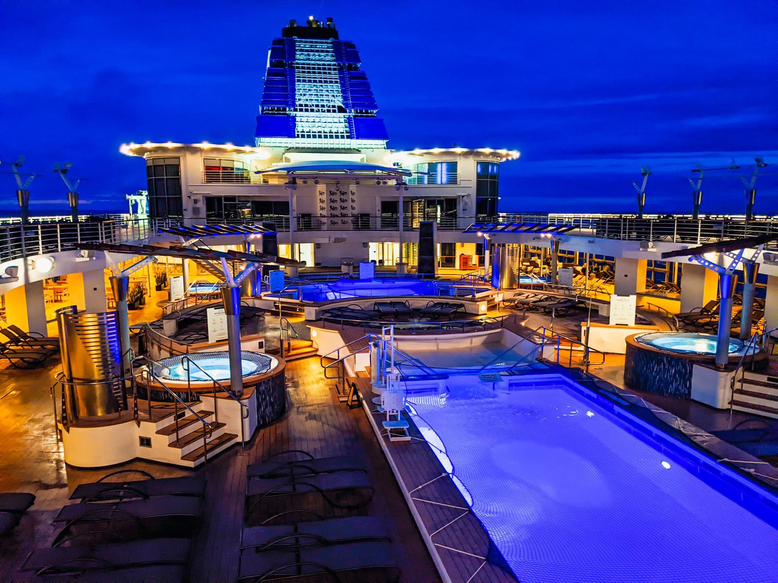 A cruise ship pool deck with lit hot tubs and loungers at twilight, where many cruise hidden costs start with a drink at the bar