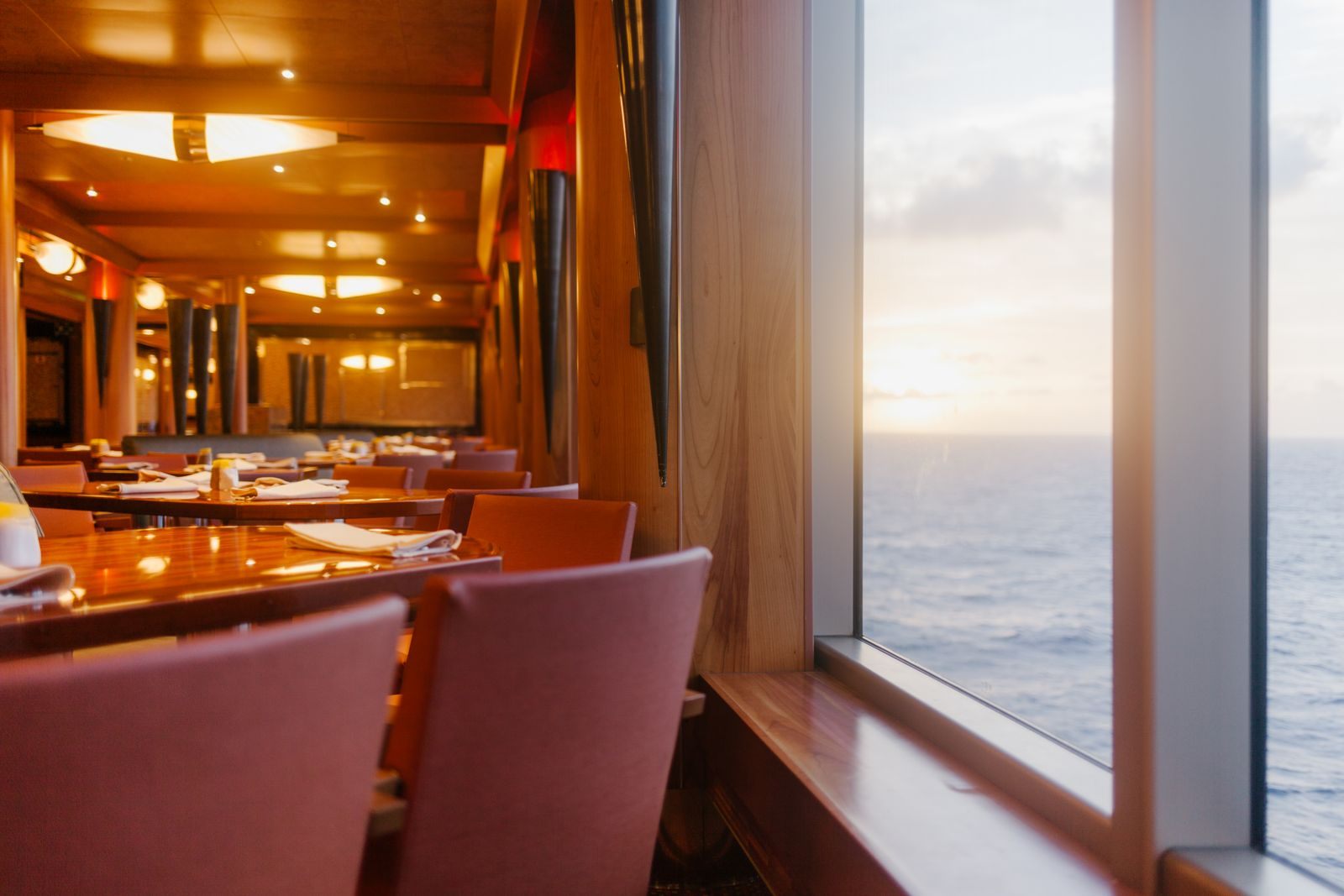 An empty wood-paneled ship dining room with tables by a window looking out to sea at sunset, the dining staff&rsquo;s domain in any cruise tipping guide